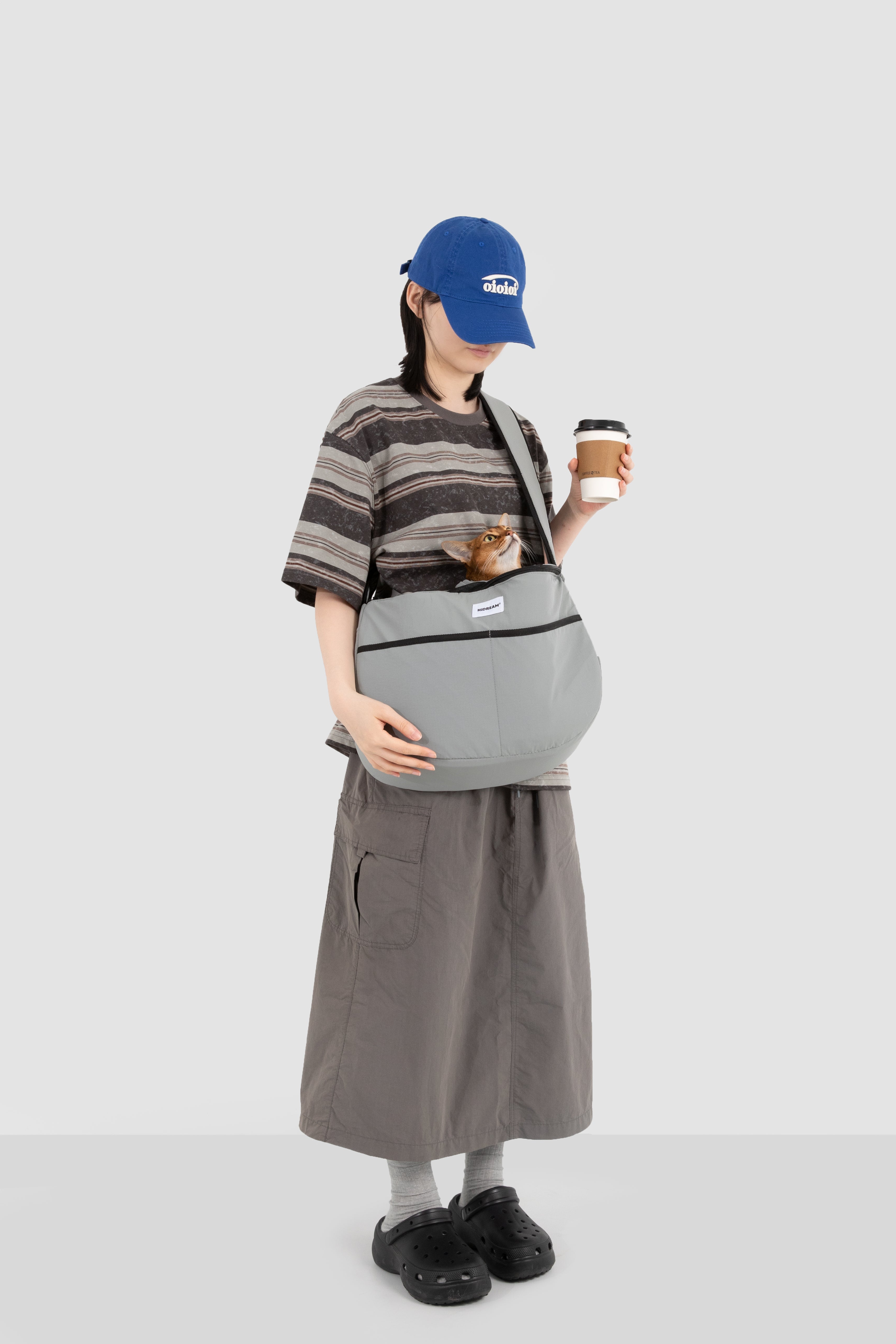 Grey HiDREAM Crossbody Pet Carrier Tote worn by a woman holding coffee, with a small dog inside. Perfect for stylish, hands-free pet transport.