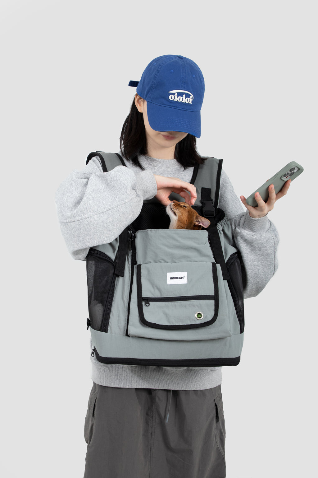 Gray HiDREAM Mountain Series Chest Pet Tote worn by woman holding phone, with small dog peeking out of breathable mesh top.