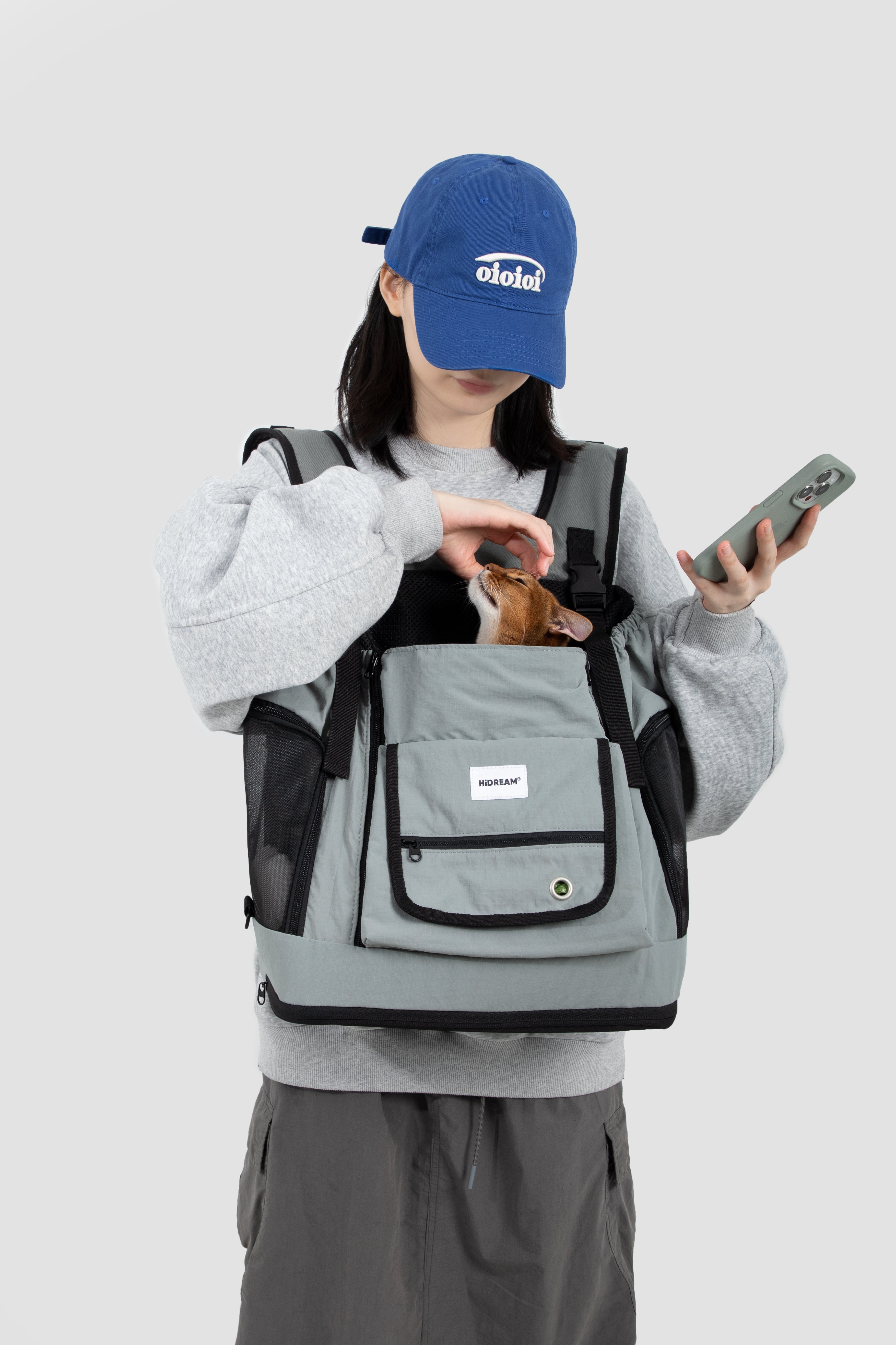 Gray HiDREAM Mountain Series Chest Pet Tote worn by woman holding phone, with small dog peeking out of breathable mesh top.