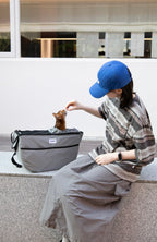 Woman feeding a cat in grey HiDREAM Crossbody Pet Carrier Tote on a bench. Comfortable and secure for cats during outdoor breaks.