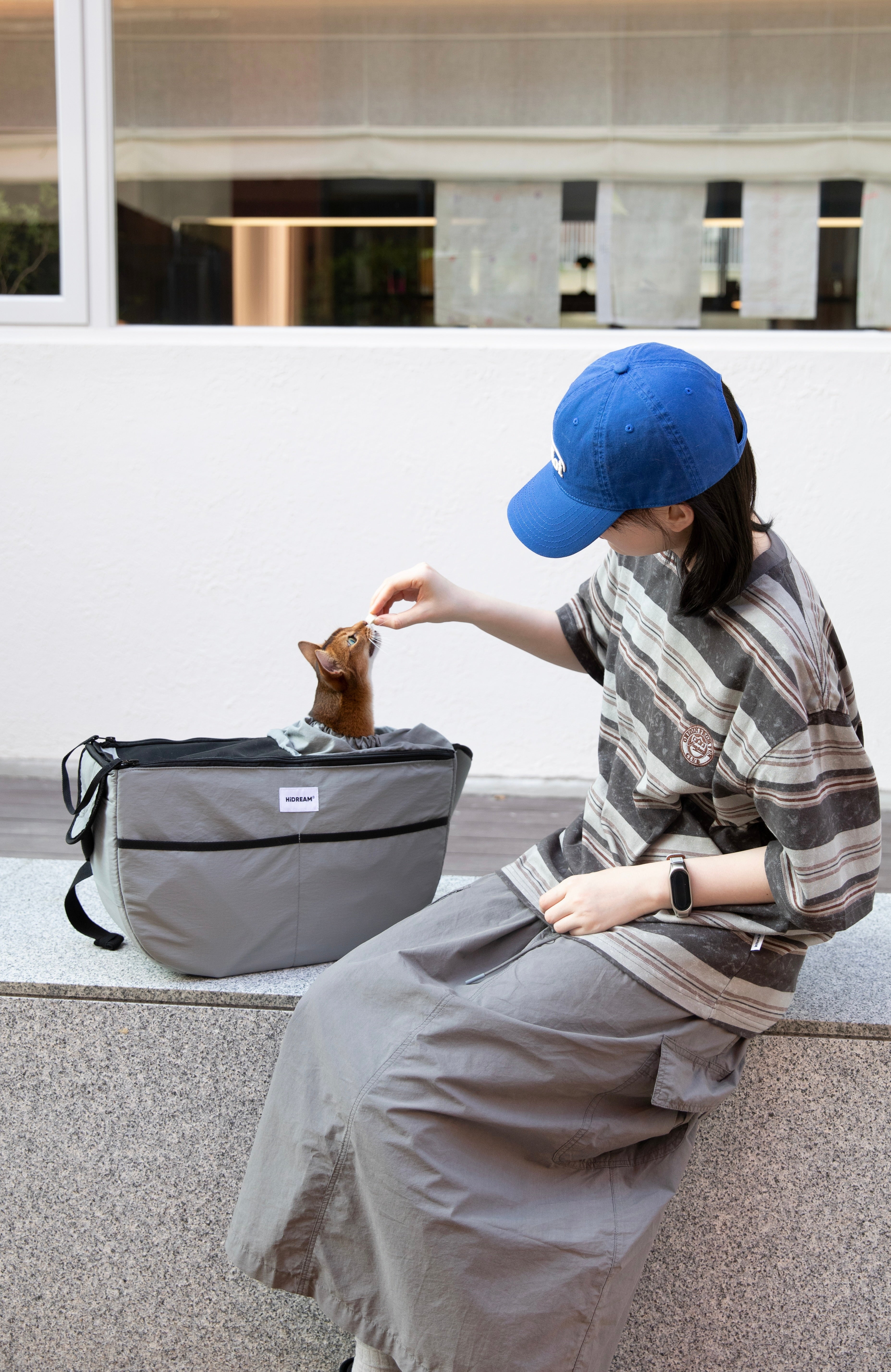 Woman feeding a cat in grey HiDREAM Crossbody Pet Carrier Tote on a bench. Comfortable and secure for cats during outdoor breaks.